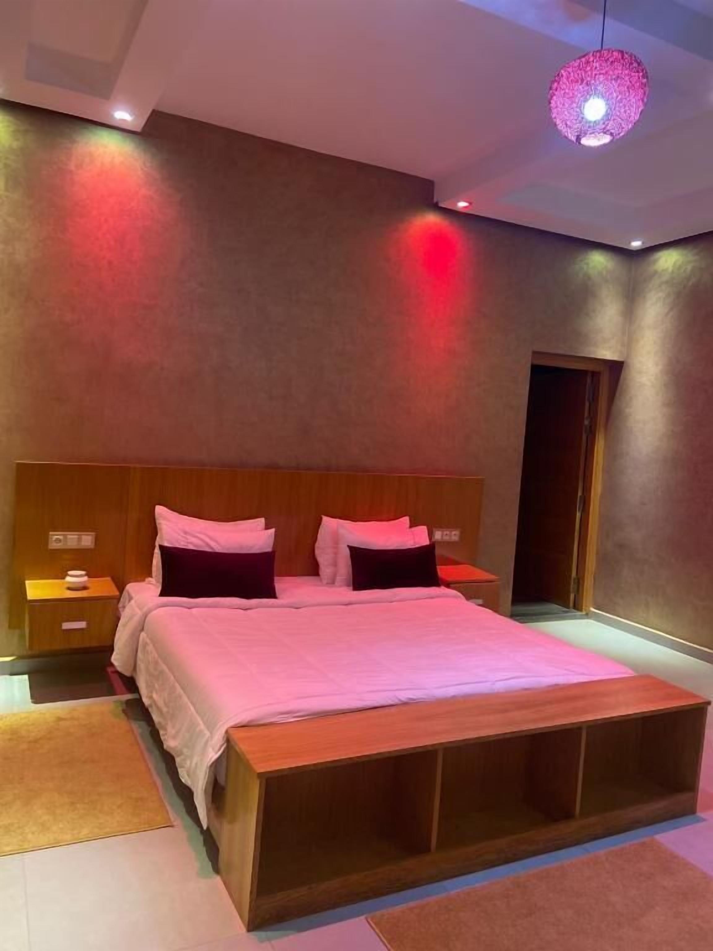 A modern bedroom with a wooden headboard, a large bed with white and dark pillows, a wooden nightstand, and a unique pink pendant light.