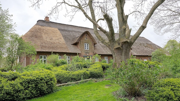 Une maison traditionnelle en briques, avec un toit de chaume, entourée de haies bien entretenues et d’une pelouse.