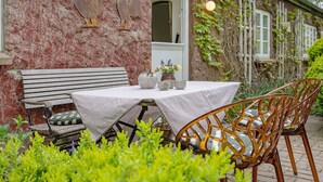 Un espace extérieur aménagé dans un style rustique, avec un banc et des chaises, une table recouverte d’une nappe et un coq décoratif accroché au mur.