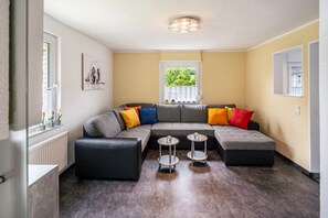 Un salon moderne avec un canapé d’angle, deux tables d’appoint, un radiateur et une fenêtre donnant sur un paysage verdoyant.
