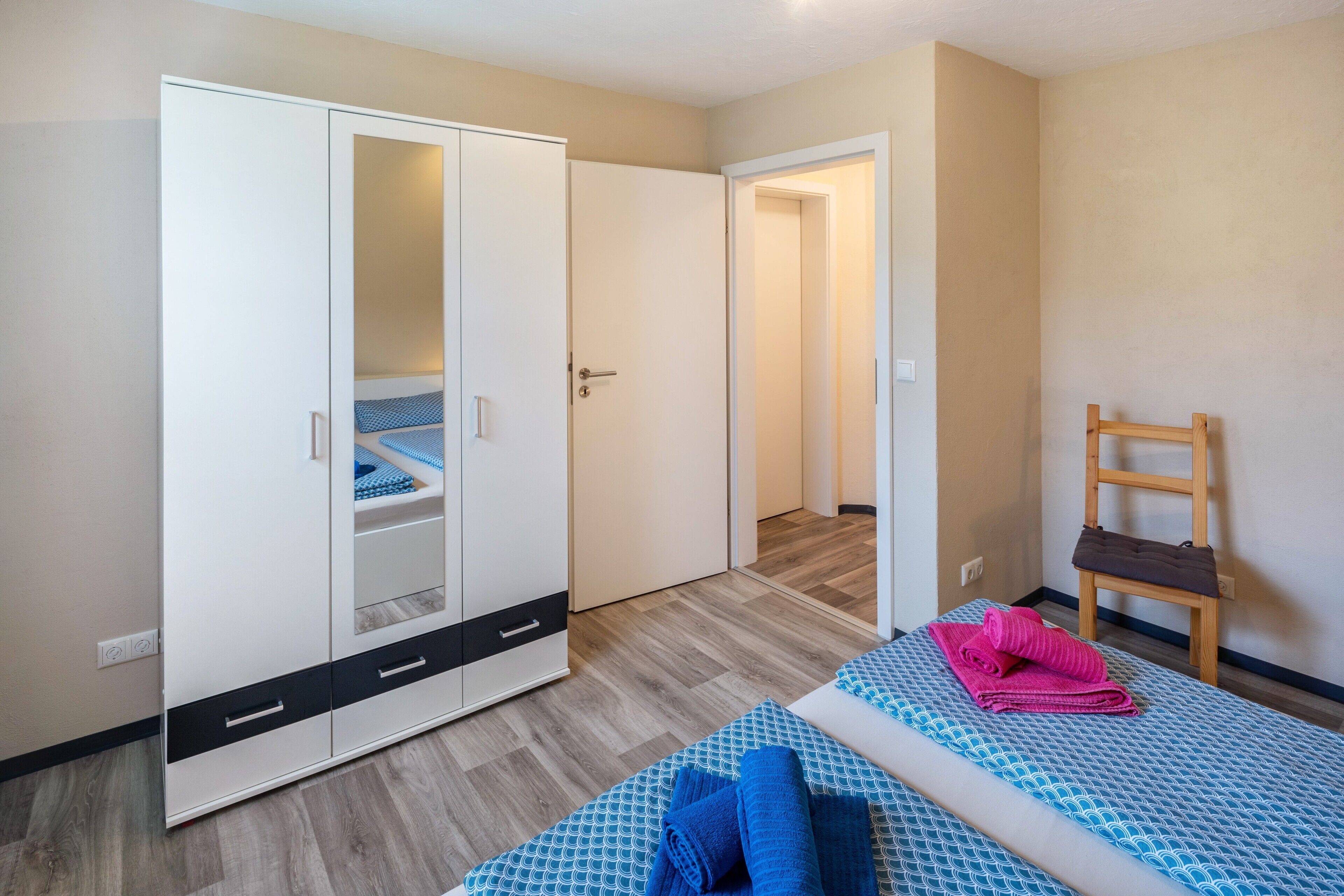 A bedroom with a bed, a wardrobe, a chair, and a door leading to another room.