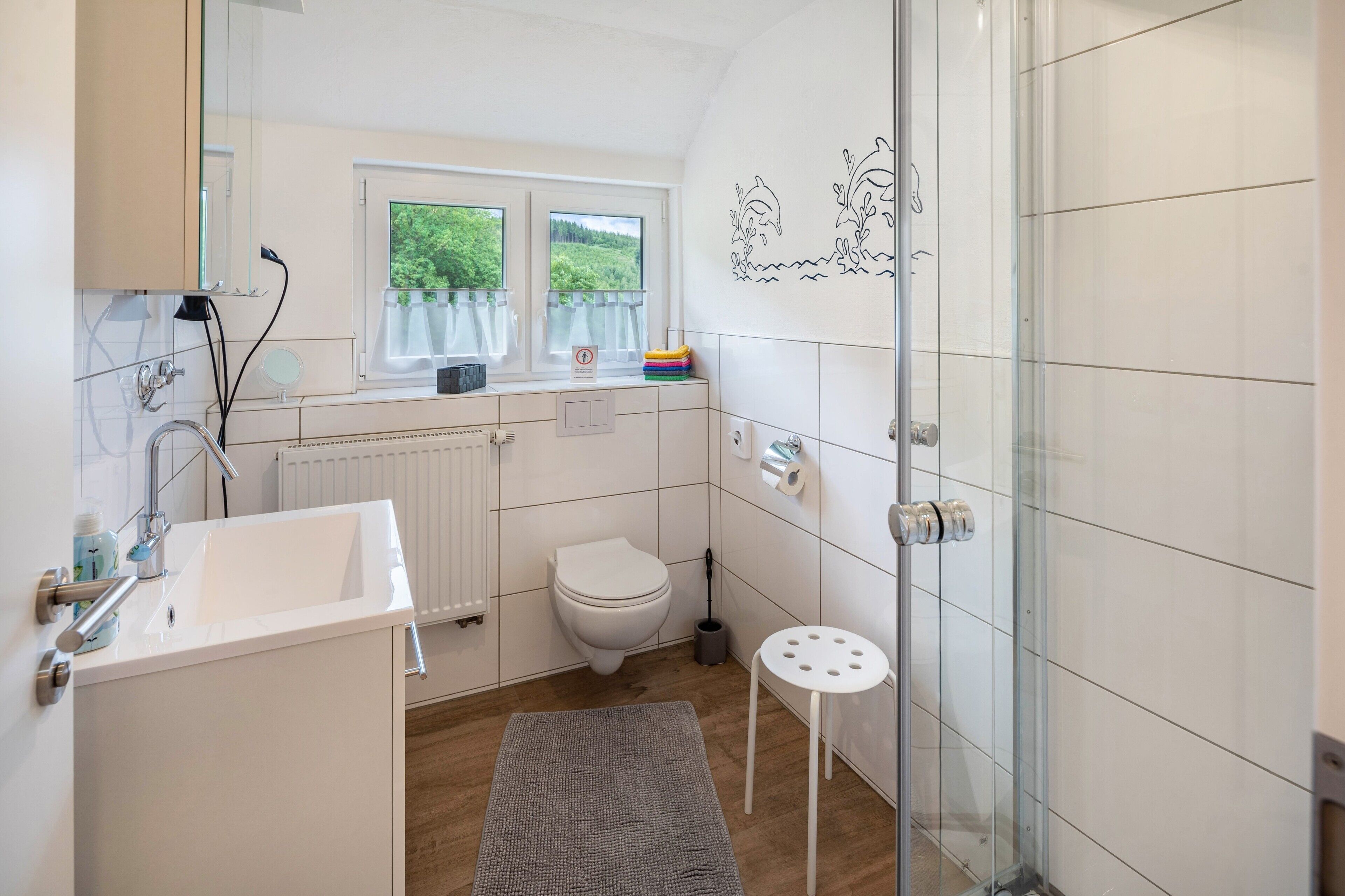 Ein modernes Badezimmer mit einem weißen Waschbecken, einer Toilette und einer Dusche. Ein Fenster bietet Blick auf viel Grün draußen.
