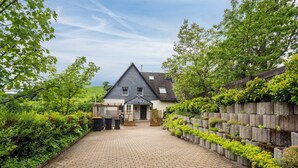 A house with a paved driveway leading to a wooden gate, surrounded by lush greenery and a stone wall with plants. - Holiday Home 'Maag, 102qm' with Private Terrace and Wi-Fi (Attendorn)