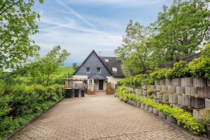 Une maison avec une allée pavée menant à un portail en bois, entourée d’une végétation luxuriante et d’un mur de pierre orné de plantes.