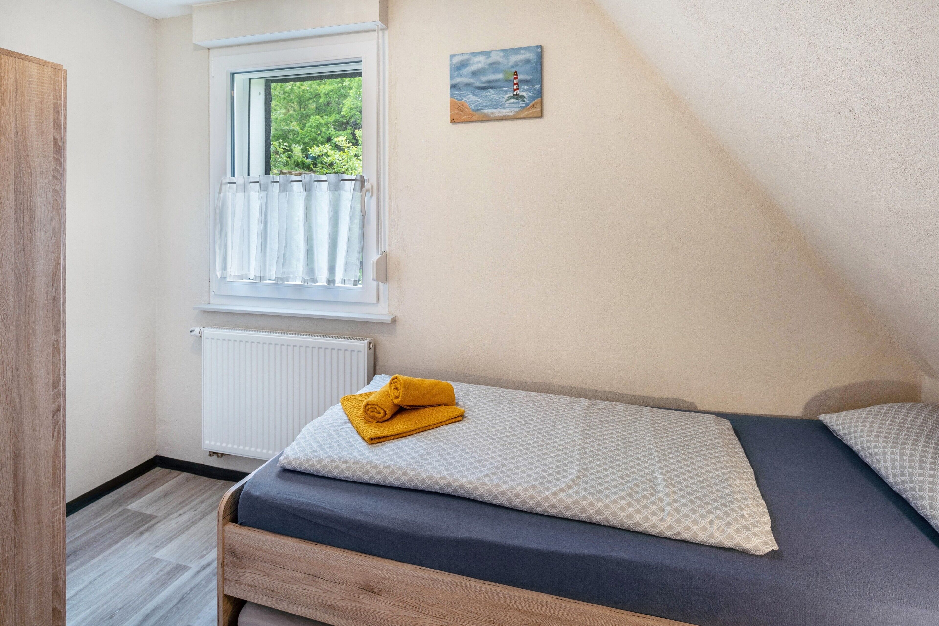 Une chambre avec une fenêtre, un tableau, un lit avec une serviette enroulée et un radiateur.