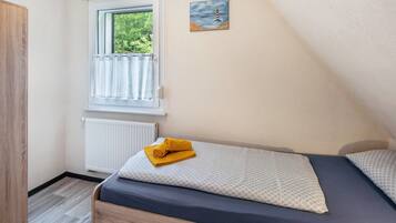 Une chambre avec une fenêtre, un tableau, un lit avec une serviette enroulée et un radiateur.