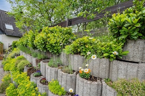 Un jardin avec différentes plantes et fleurs, une clôture en bois et une maison en arrière-plan.