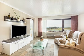 Una sala de estar moderna con un televisor de pantalla plana, una mesa de centro de vidrio, un sofá color beige y vistas al exterior a través de amplios ventanales.