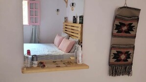 A bedroom with a bed, a wooden headboard, a mirror, a shelf with toiletries, and a hanging blanket. - Sea shy apartment (Dahab)