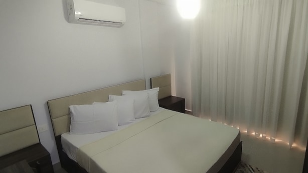 A hotel room with a bed, white curtains, a bedside table, and an air conditioning unit.