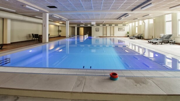 Une grande piscine intérieure avec un plafond carrelé, des chaises longues et un petit objet rouge placé au bord du bassin.