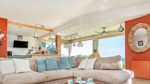 Una sala de estar moderna con un amplio sofá seccional color beige, una mesa de centro de madera con teteras y tazas, y una cocina al fondo.