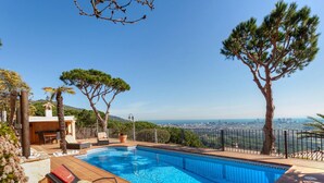 Piscina privada con vistas a un horizonte urbano, rodeada de árboles y un patio con sillones de descanso.