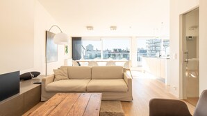 A modern living room with a sofa, a wooden coffee table, and a TV. There is a large window with a city view.