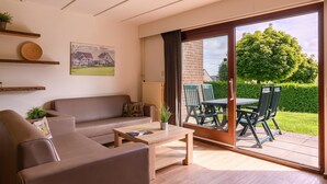 A modern living room with a sofa, coffee table, and a sliding glass door leading to an outdoor dining area with chairs and a table. - Bungalow in Reevallis near Scenic Hills (Vijlen)