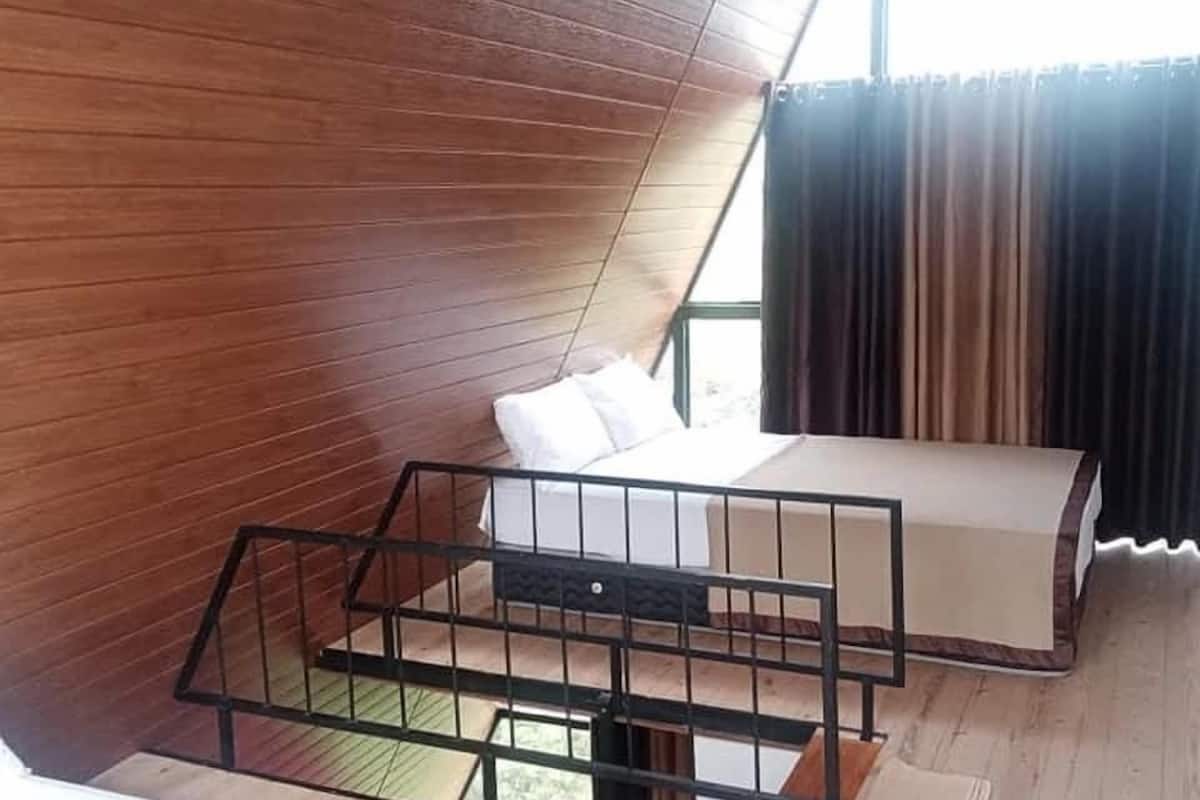 A room with a sloped ceiling, wooden walls, a bed with a mattress and pillows, a small table, and a window with curtains.