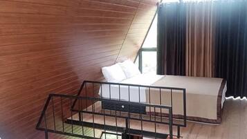 A room with a sloped ceiling, wooden walls, a bed with a mattress and pillows, a small table, and a window with curtains.