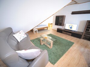 Un salon moderne avec un canapé, une table basse en verre, un téléviseur et un petit lit placé sous un plafond en pente.