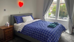 A neatly made bed with a blue and white checkered bedspread, a wooden nightstand with a tissue box, and a red heart-shaped light on the wall. - Victorian terrace house (Edinburgh)