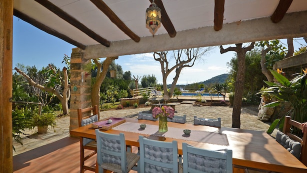 An outdoor patio with a wooden table and chairs, surrounded by potted plants and a view of a swimming pool and mountains. - Rafal baix - idyllic country house with private pool (Son Servera)