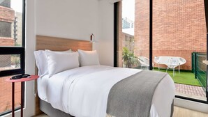 Quarto de hotel moderno com uma cama grande, uma mesinha auxiliar, uma janela com vista para um edifício de tijolos e uma varanda com área de estar externa.