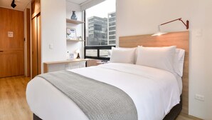 Quarto de hotel moderno com uma cama grande, cabeceira de madeira, mesa de cabeceira, janela com vista para a cidade e um guarda-roupa embutido.