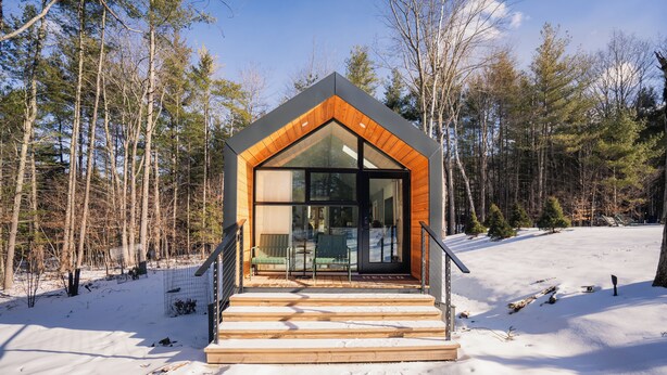 A modern cabin with a wooden exterior and large glass windows, surrounded by a snowy forest.