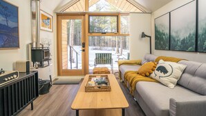 A cozy living room with a wooden table, a sofa with cushions, a fireplace, and a large window offering a view of the outdoors.
