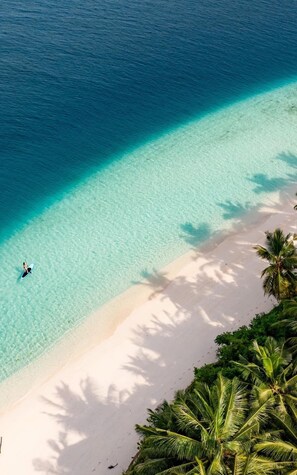 Luchtbeeld van een strand met helder turquoise water, een persoon op een paddleboard en toekijkende palmbomen.