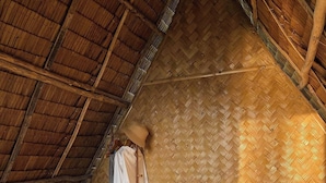 Ein gemütliches Schlafzimmer mit einem Strohdach, einem Bett, einem Nachttisch, einem Ventilator und einem Kleiderständer.