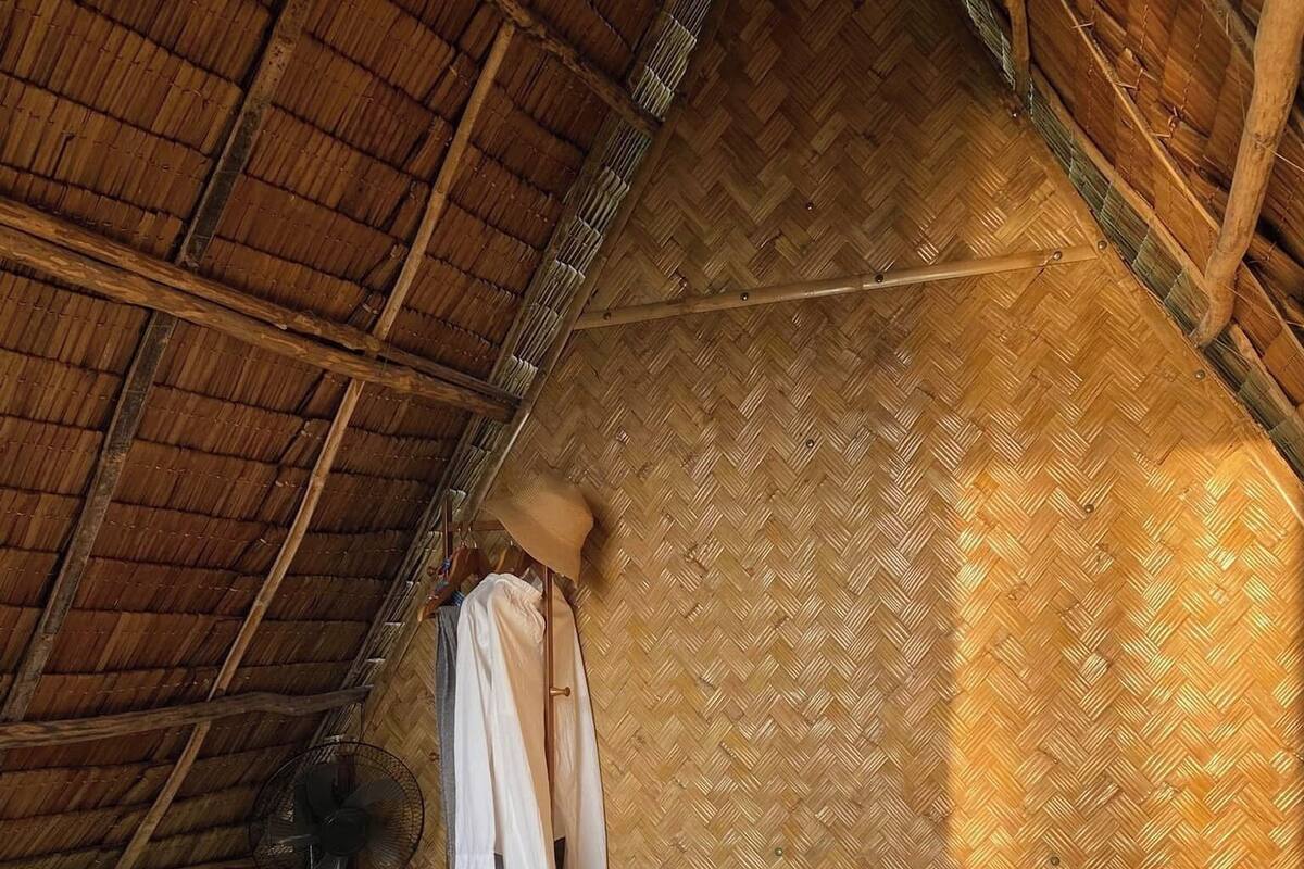 Een gezellige slaapkamer met rieten dak, een bed, nachtkastje, ventilator en kledingrek.