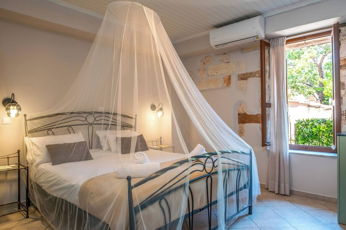 Una cama bien hecha con mosquitero, una lámpara de noche y vistas al follaje exterior.