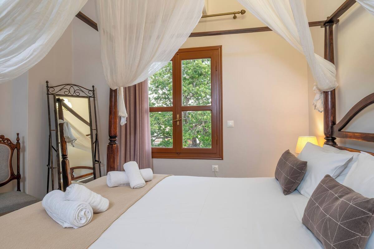 Un dormitorio con una cama grande, una ventana con vistas a árboles, un espejo y una mesita de noche con toallas.