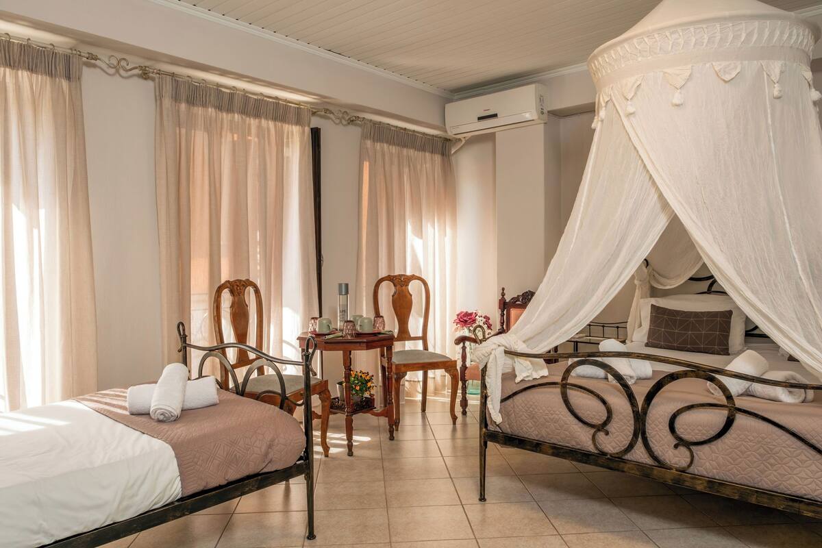 Habitación de hotel con dos camas individuales, una silla de madera, una mesita con un jarrón de flores y un ventilador de techo.
