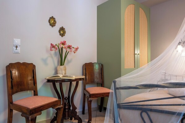 Un dormitorio con una cama cubierta por una mosquitera, dos sillas de madera, una mesita con un libro y flores, y un interruptor de luz en la pared.