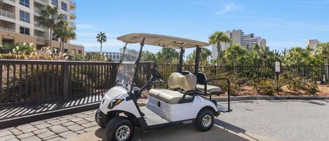 A golf cart parked on a paved area with a fence and palm trees in the background.