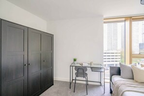 A bedroom with a bed, a desk, a chair, a wardrobe, and a window with a city view. - Central Birmingham 1BR Flat with Comfort & Style (Birmingham)