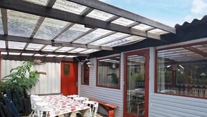 Ein überdachter Patio-Bereich mit Tisch und Stühlen, eine rote Tür und ein Fenster mit Blick auf den Garten.