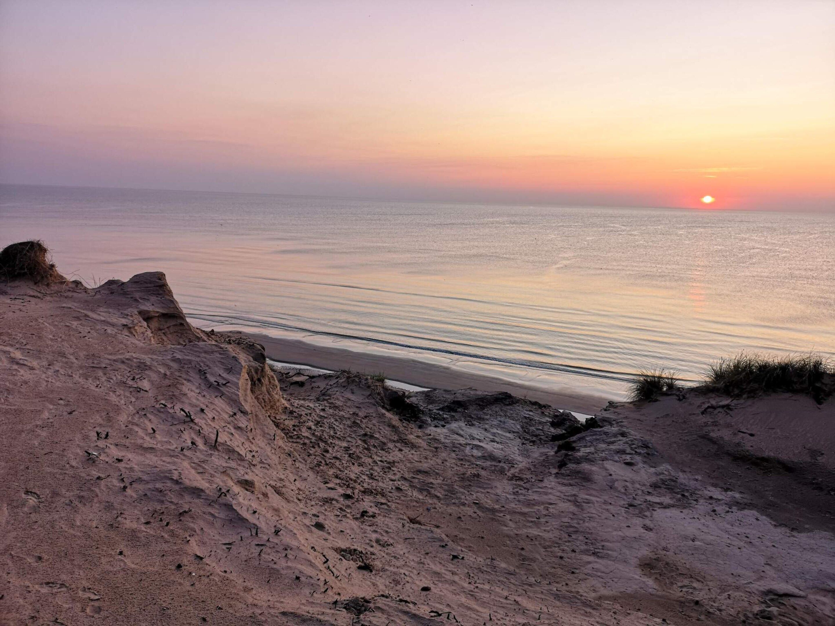 Eine Küstenlandschaft mit Sonnenuntergang über dem Meer, sandigem Gelände und Felsformationen.