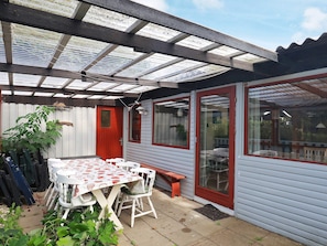 A covered patio area with a table and chairs, a red door, and a window with a view of the garden. - 6 Person Holiday Home in Lokken (Lokken)