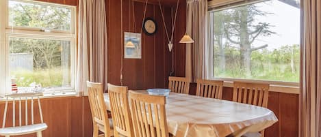 A dining area with a wooden table and chairs, large windows with curtains, a clock on the wall, and a pendant light.