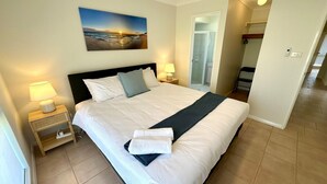 Une chambre d’hôtel avec un grand lit, une table de chevet avec une lampe, un tableau représentant une scène de plage et une salle de bain visible à travers une porte ouverte.