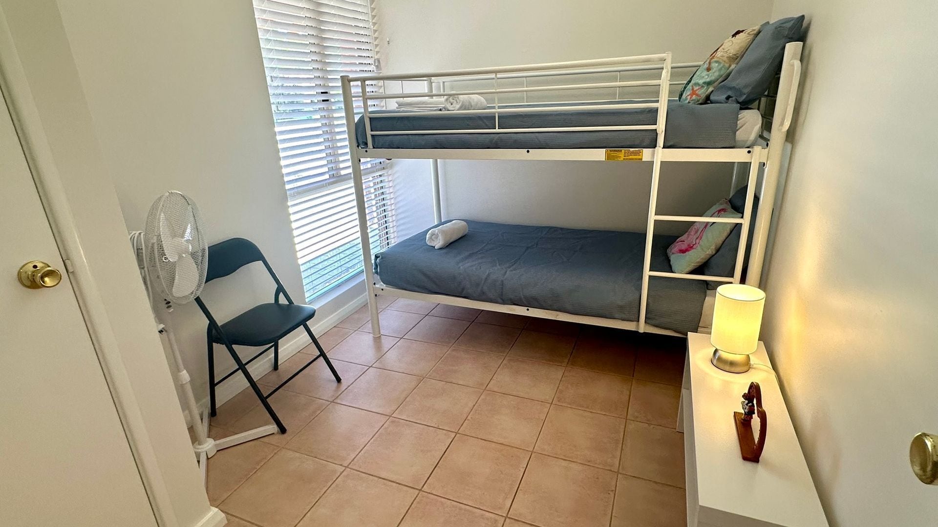 Een kleine, compacte kamer met een stapelbed, een stoel, een ventilator en een nachtkastje met een lamp.