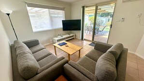Un salon avec deux canapés gris, une table basse en bois, un téléviseur à écran plat et une porte-fenêtre coulissante donnant sur une terrasse extérieure.