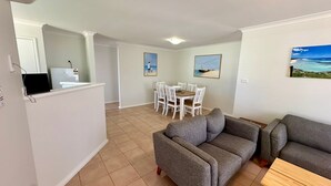 Un salon avec un canapé gris, un coin repas avec une table et des chaises blanches, et une cuisine avec un comptoir et un téléviseur.