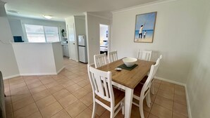 Un coin repas avec une table en bois et des chaises blanches, un réfrigérateur blanc et un tableau représentant une scène de plage.