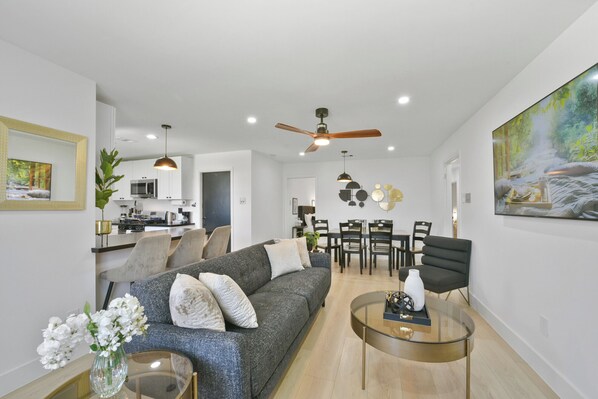 Ein modernes Wohnzimmer mit einem grauen Sofa, einem Glastisch und einem Essbereich mit einer Chandelier.