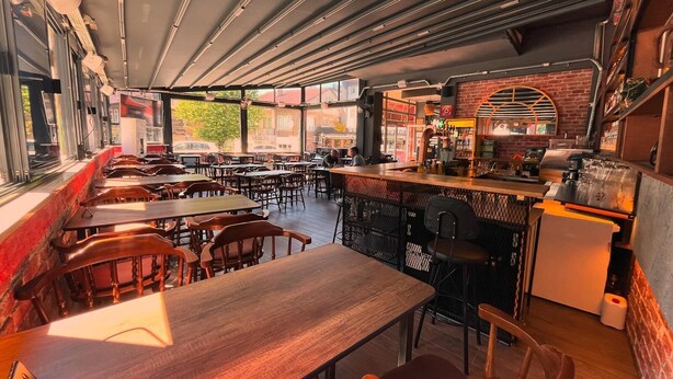Un restaurante con mesas y sillas de madera, una zona de bar con una parrilla, y grandes ventanales que ofrecen vistas a la calle.
