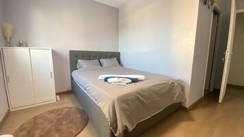 Un lit bien fait, avec une tête de lit grise, une armoire blanche et un petit tapis rond sur un plancher en bois.
