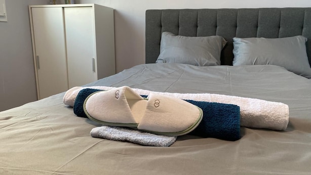 Un lit bien fait, avec une tête de lit grise, une paire de chaussons et une serviette enroulée posée sur un couvre-lit beige.